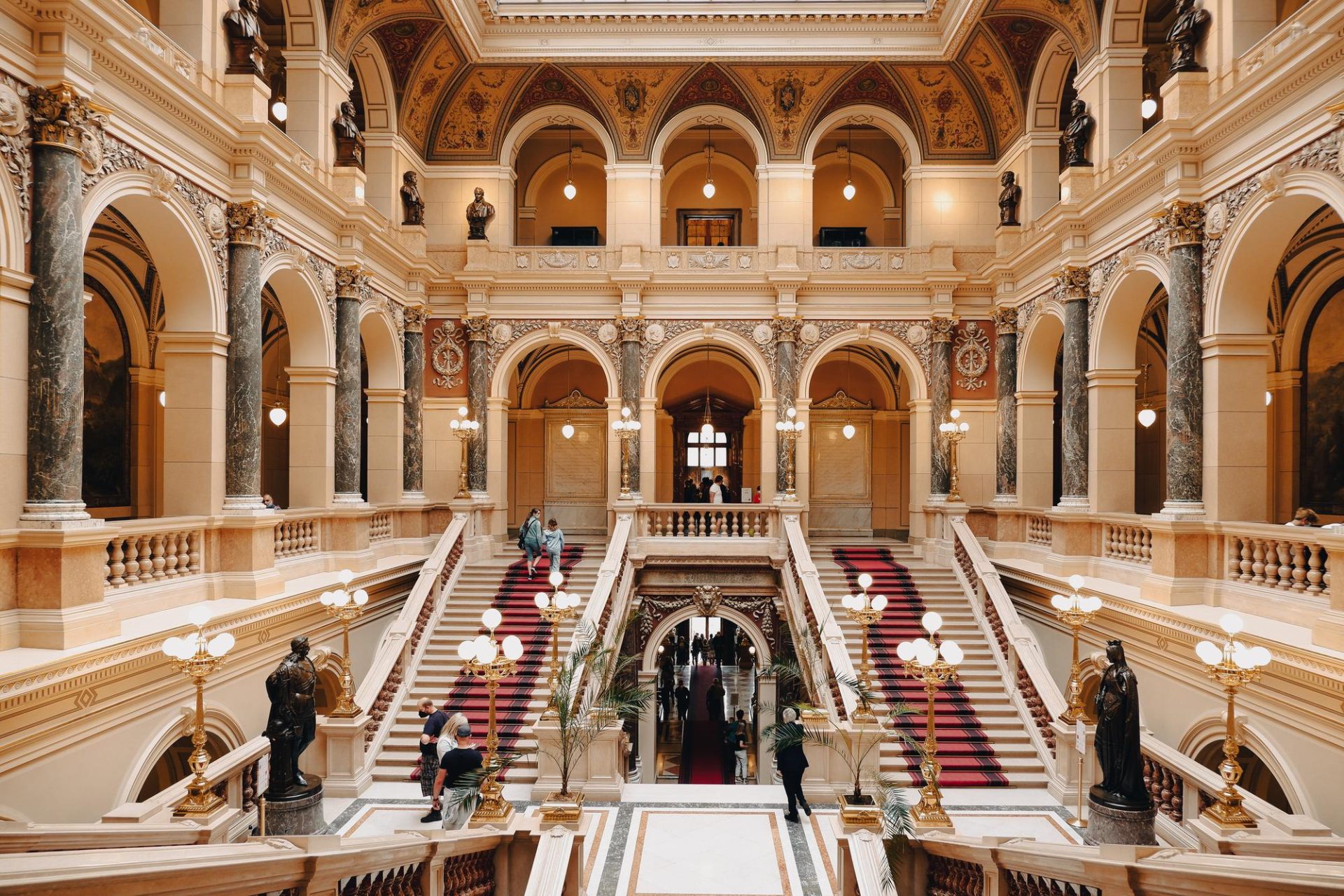 Ornate historic interior with grand staircases, arches, and people in motion.